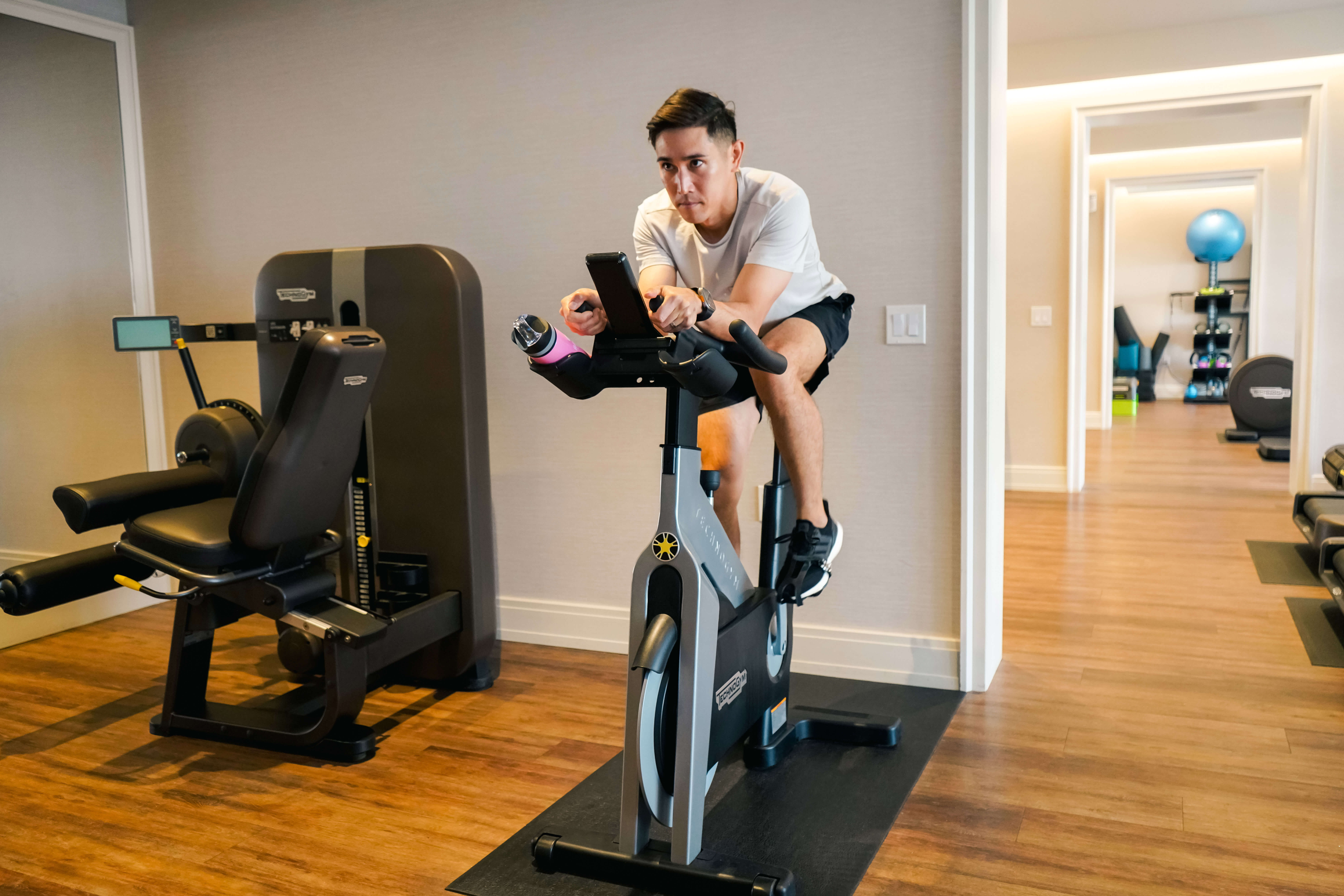 白いシャツと黒いショートパンツ姿の男性が、屋内でスマートフォンを見ながらエアロバイクをこいでいます。室内にはトレーニング機器と木の床があります。