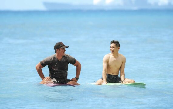 海でサーフボードに座り、笑顔で語り合う2人の男性。空と海は鮮やかな青で、背景にはぼやけた船が見えます。